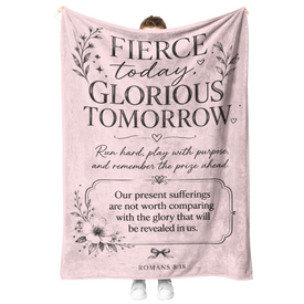 A person holds up a large pink fleece blanket with black printed inspirational text and floral illustrations. The blanket reads, 'Fierce today, glorious tomorrow' and includes a Bible verse from Romans 8:25-28.