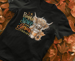 Black t-shirt with a graphic of a highland cow holding a pumpkin spice latte and text 'Pumpkin Spice and Jesus Christ' on a fall-themed background.